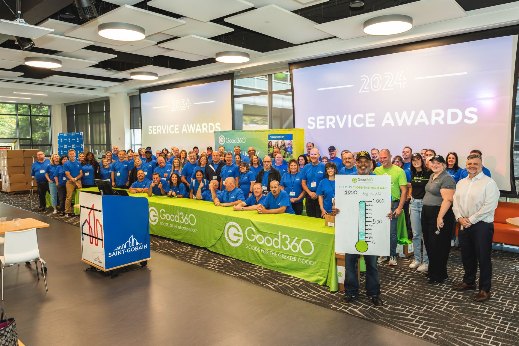 A large group of people wearing matching shirts pose behind a Good360 table