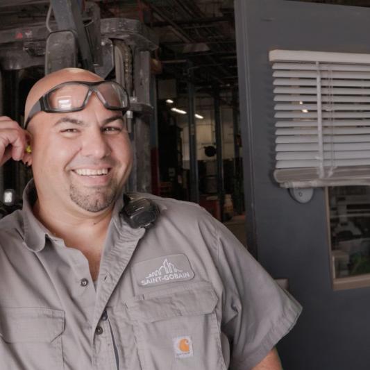 A man wearing safety goggles on the top of his head smiles in the office area of the manufacturing building.