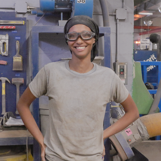 A Black woman wearing a head covering and safety goggles stands on the manufacturing floor