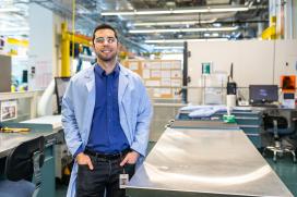 A man in a blue lab coat stands next to a metal table in a manufacturing setting. He is wearing safety goggles.