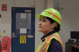 A woman wearing a yellow hard hat and safety vest stands in the manufacturing plant