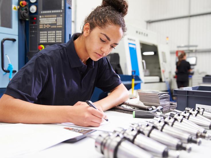 Young female design engineer working with blueprints and tools on her desk, there is manufacturing machinery and a prototyping worker in the background