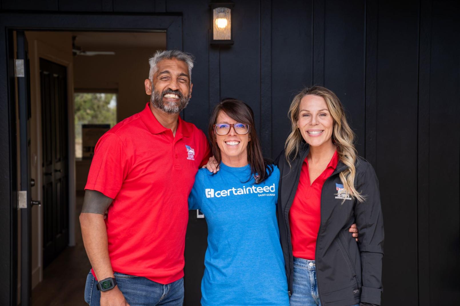 A man wearing a red shirt and his wife, also wearing a white shirt, pose with a CertainTeed employee in front of their new home