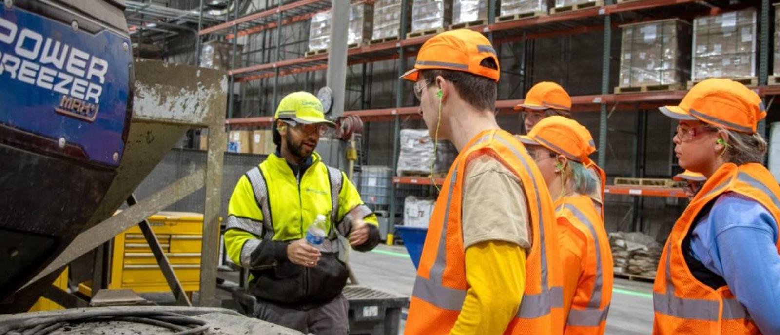 A group of trainees wearing orange high visibility vests and safety hardhats listening to an instructor during on-the-job training for entry level plant jobs.