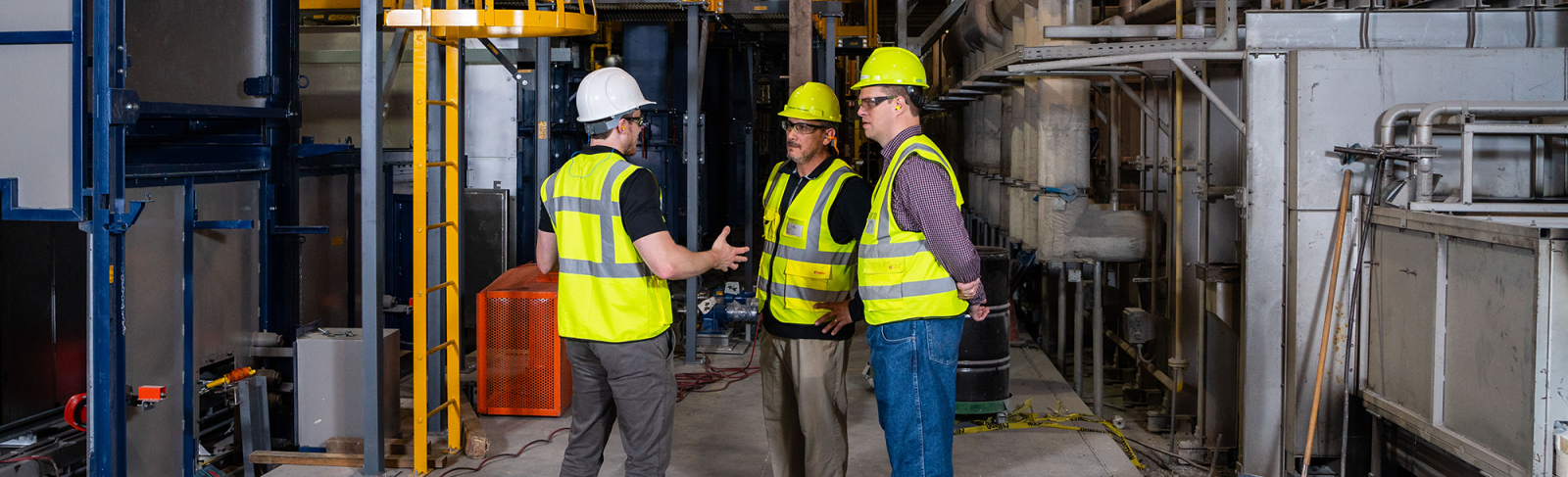 Three men in yellow safety gear are standing on the manufacturing floor