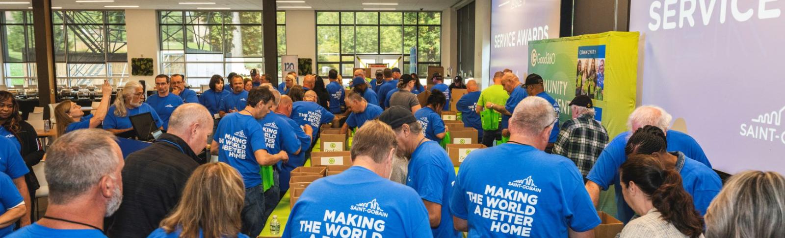 A large group of people wearing matching tshirts that say "Making The World A Better Home" are packing Good360 hygiene kits in an assembly line