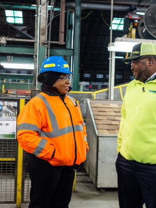 A Black man and a Black woman are standing in a factory setting. She wears a blue hard hat and orange safety jacket. He wears a yellow safety jacket. 